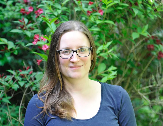 Dr Carole Minker, Health professional partner of SOVA, wearing glasses and a dark top, standing outside with green plants and pink flowers behind her.